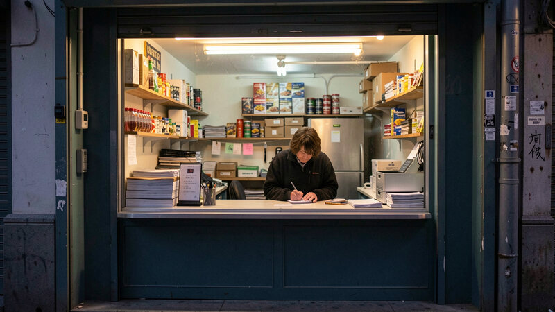 A closed shop front with the lights on inside, a staff member writing on a notepad behind the counter in the evening.
