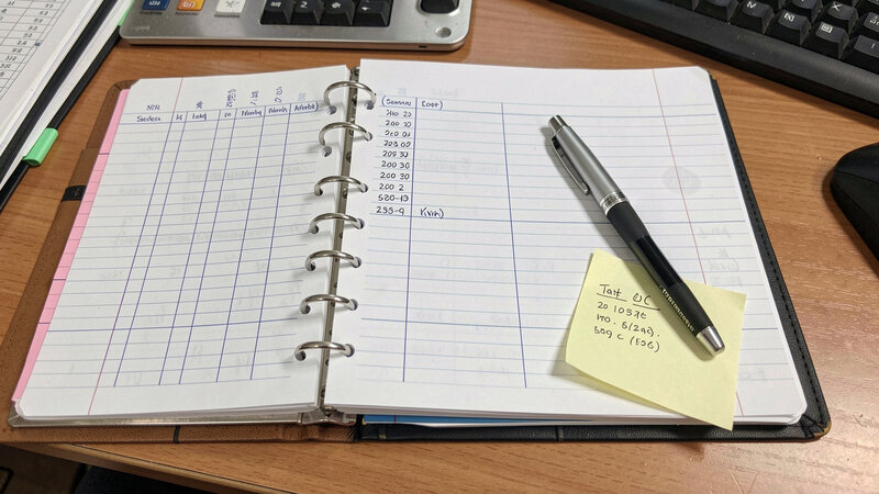 A ring binder open on a desk showing a simple handwritten log table, with a pen and a dated sticky note on the page.